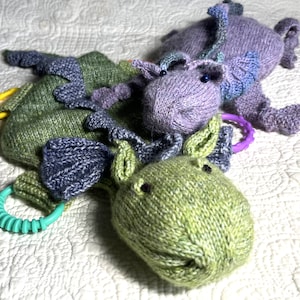 Könnte beinhalten: Drei gestrickte Plüschtiere in Form von Drachen. Die Drachen sind grün, lila und blau. Die Drachen haben große Augen und sind aus einem weichen, strukturierten Garn gefertigt.