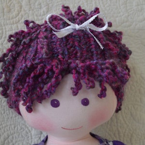 May include: A handmade doll with purple and pink yarn hair, a white bow, and a pink blush on its cheeks.