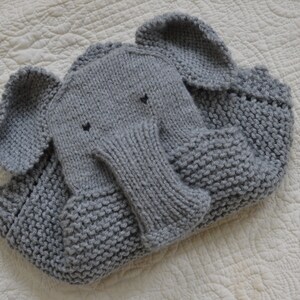 May include: A gray knitted elephant blanket with a textured surface. The blanket is folded and the elephant's trunk is visible.