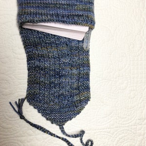 May include: A blue and green knitted book cover with a tassel. The cover is made of a soft, textured yarn and has a unique, hand-knitted design.