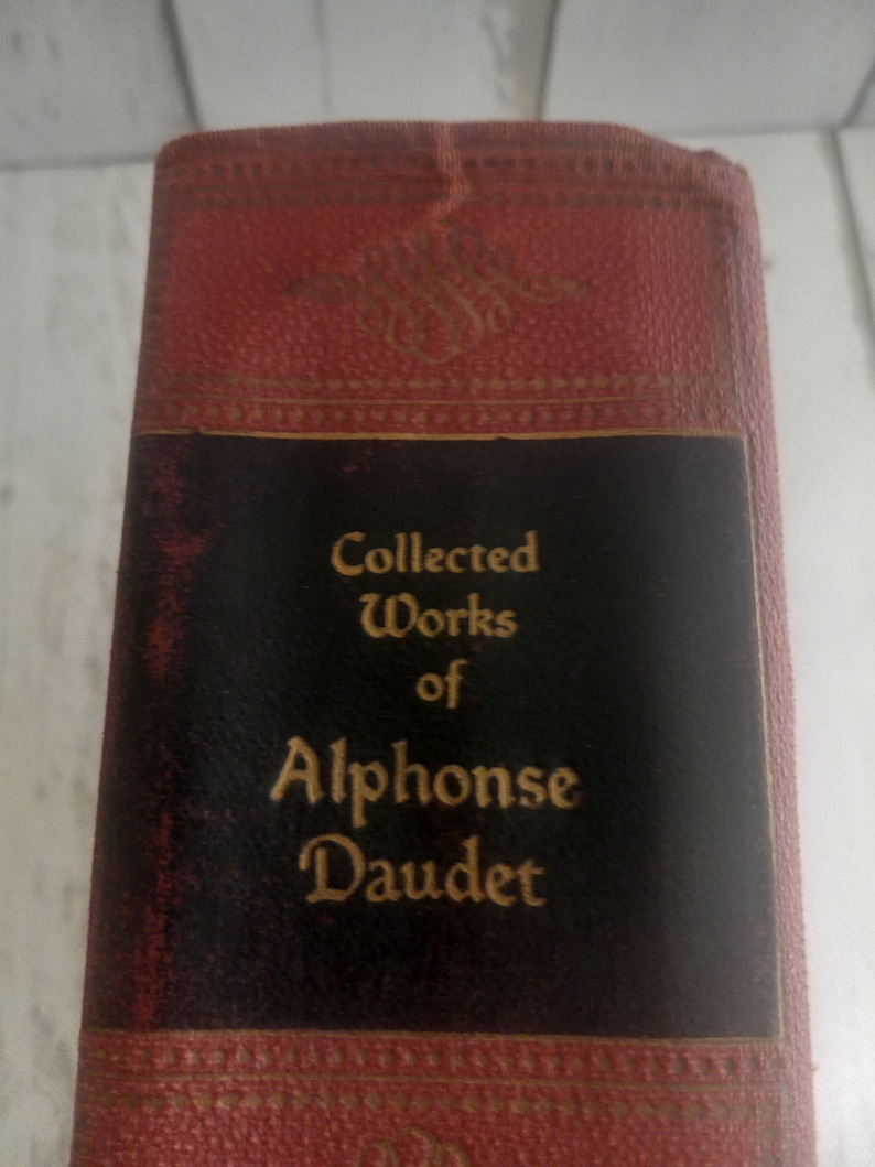 K&ouml;nnte beinhalten: Ein rotes, ledergebundenes Buch mit goldenen Lettern auf dem R&uuml;cken. Der Titel lautet "Gesammelte Werke von Alphonse Daudet".