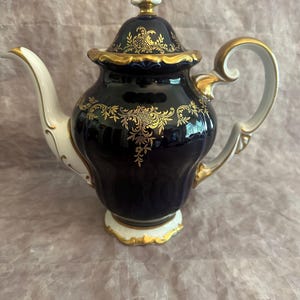 May include: A navy blue porcelain teapot with gold filigree detailing. The teapot features a white handle and spout, with gold trim. The lid is navy blue with gold accents and a white finial. A decorative piece for tea parties.
