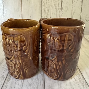 May include: Two brown ceramic mugs with ornate, raised floral and scrollwork designs. The mugs have a handle and a slightly flared rim. The background is a light-colored wooden surface.