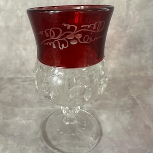 May include: A vintage clear glass goblet with a ruby red rim. The rim features a white etched floral design. The lower portion of the goblet has a textured pattern. The goblet sits on a clear, round base.