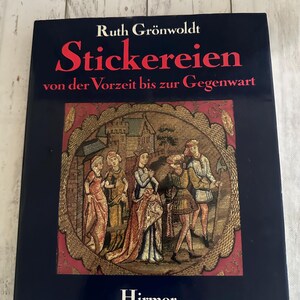 May include: A book titled "Stickereien" by Ruth Grönwoldt, with text in German. The cover is dark blue with red and white text. It features an illustration of people in medieval clothing within a decorative frame. The publisher's name, Hirmer, is at the bottom.
