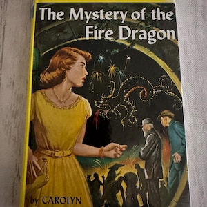 May include: A vintage Nancy Drew Mystery Stories book titled "The Mystery of the Fire Dragon." The cover features a woman in a yellow dress, fireworks, and illustrations of people. The author is Carolyn Keene.