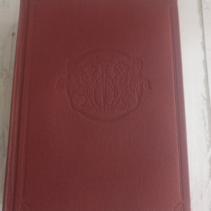K&ouml;nnte beinhalten: Ein burgunderfarbenes Hardcover-Buch mit einem dekorativen, gepr&auml;gten Design auf dem vorderen Umschlag. Das Design zeigt einen runden Rahmen mit einem stilisierten Schwert und zwei V&ouml;geln.