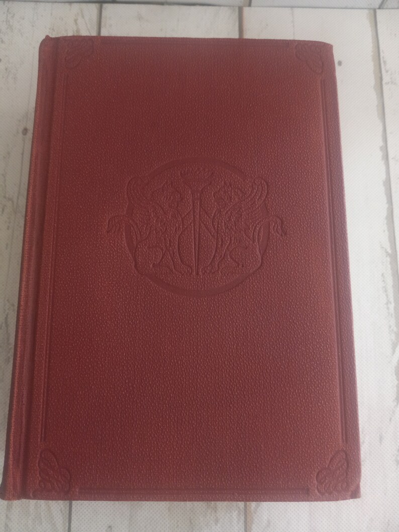 K&ouml;nnte beinhalten: Ein burgunderfarbenes Hardcover-Buch mit einem dekorativen, gepr&auml;gten Design auf dem vorderen Umschlag. Das Design zeigt einen runden Rahmen mit einem stilisierten Schwert und zwei V&ouml;geln.