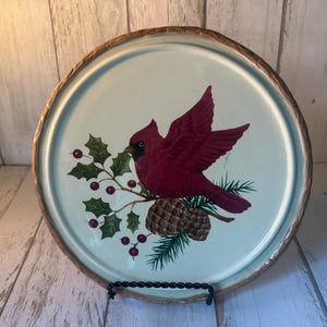 May include: A round decorative plate with a cream-colored center and a textured brown rim. The center features a vibrant red cardinal bird perched on a branch with green holly leaves, red berries, and two brown pinecones against a light background.