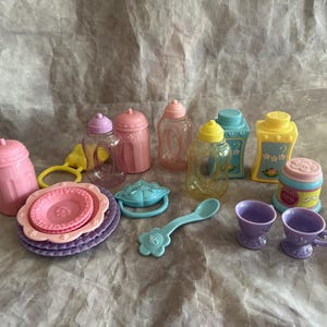 May include: A collection of colorful toy baby bottles, plates, cups, and accessories. The set includes pink, purple, yellow, and blue items, including a rattle, a spoon, and a stack of plates. The toys are made of plastic.