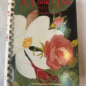 May include: A spiral-bound cookbook titled A Cook's Tour of Shreveport Favorite Southern Recipes by the Junior League of Shreveport. The cover features a large white magnolia flower, a red rose, and a crawfish against a dark background.