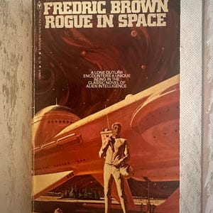 May include: Vintage paperback book cover for "Fredric Brown: Rogue in Space." The cover features a retro sci-fi illustration of a man in a spacesuit standing in front of a spaceship with a red and orange space background. The book title is prominently displayed.