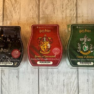 May include: Three Scentsy wax bars in clear plastic packaging. The left bar is black with a castle illustration, the center is red with a Gryffindor crest, and the right is green with a Slytherin crest. Each bar is labeled Harry Potter and 2.6 oz.