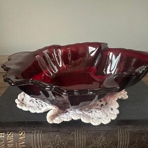 May include: A ruby red glass bowl with a scalloped edge. The bowl is resting on a white crocheted doily and a dark book. The bowl has a glossy finish and a unique, flower-like shape.