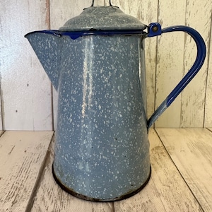 May include: A vintage blue and white speckled enamel coffee pot with a blue rim and handle. The pot has a lid with a black handle.