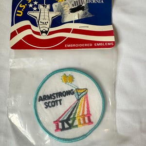 May include: A circular embroidered patch in a clear plastic bag with a U.S. Space Camp header card. The patch features a white background, a light blue border, the text ARMSTRONG SCOTT, two yellow stars, and a colorful rainbow-striped structure.