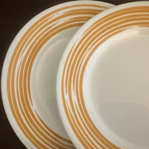 May include: Two round white ceramic plates featuring a series of concentric golden-yellow rings along the outer rim. The plates have a smooth, glossy finish and a clean, circular design that highlights the contrast between the white and yellow colors.