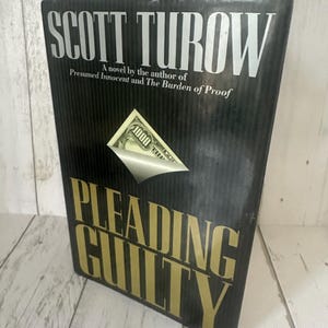 May include: A hardcover book titled "Pleading Guilty" by Scott Turow. The book has a black cover with gold lettering and a graphic of a $1000 bill. The author is also of "Presumed Innocent" and "The Burden of Proof".