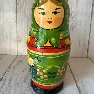 May include: A wooden nesting doll featuring a painted face with dark hair, wearing a green headscarf. The body is decorated with red, yellow, and green bands, and a floral pattern with small berries. The doll has a black base and shows signs of wear.