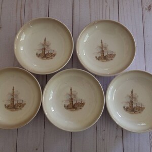 May include: Set of five cream-colored ceramic bowls with a brown design of a building with a tall tower. The bowls are arranged in a circle on a light wood surface.