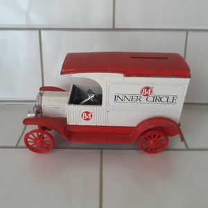 May include: A vintage toy truck in red, white, and silver. The truck features a red roof, wheels, and chassis, with a white body. The text "84 INNER CIRCLE" is printed on the side. The toy has a slot on top, suggesting it's a bank.