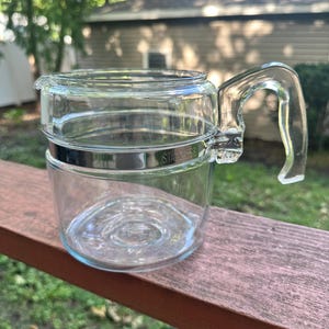 May include: A clear glass carafe featuring a polished stainless steel band around the center with the word STAINLESS embossed on it. The vessel includes a transparent glass handle attached to the metal band and a small pouring spout at the rim.