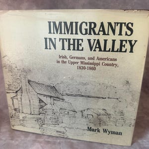 May include: A hardcover book titled Immigrants in the Valley: Irish, Germans, and Americans in the Upper Mississippi Country, 1830-1860 by Mark Wyman. The off-white cover features a black ink sketch of a rural landscape with small wooden cabins.