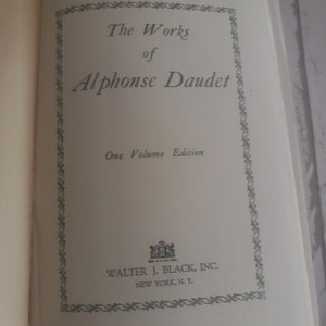 K&ouml;nnte beinhalten: Ein Buchcover mit einem dekorativen Rahmen und dem Titel "The Works of Alphonse Daudet" in schwarzer Schrift. Der Untertitel lautet "One Volume Edition". Der Verlag ist "Walter J. Black, Inc. New York, N.Y."