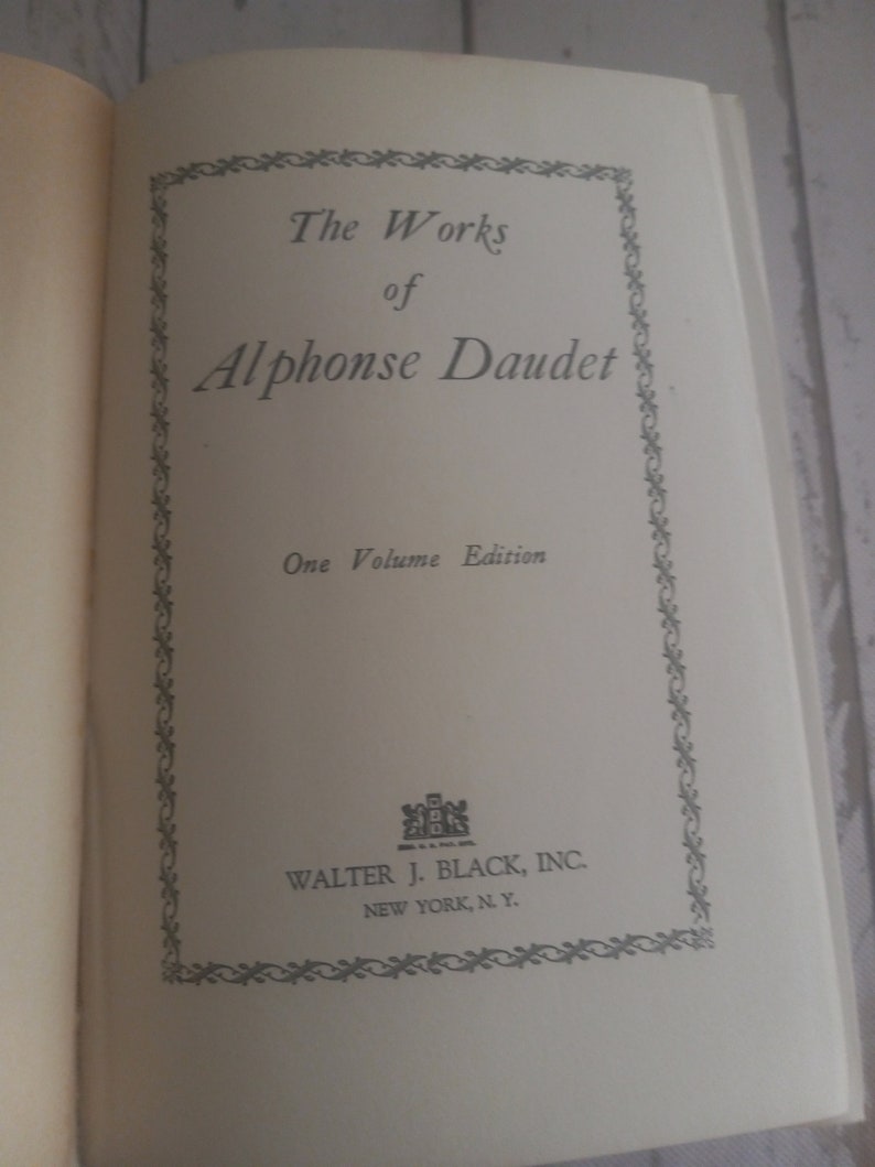 K&ouml;nnte beinhalten: Ein Buchcover mit einem dekorativen Rahmen und dem Titel "The Works of Alphonse Daudet" in schwarzer Schrift. Der Untertitel lautet "One Volume Edition". Der Verlag ist "Walter J. Black, Inc. New York, N.Y."