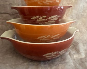 May include: A set of four nested mixing bowls with handles, featuring a repeating white wheat stalk pattern. The bowls alternate between shades of orange and reddish-brown, with each bowl displaying the same wheat design on the exterior surface.