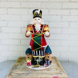 May include: A ceramic nutcracker figurine of a soldier holding a drum. The nutcracker is wearing a red and gold uniform with a black hat and white beard. The figurine is standing on a base with a small castle and presents.