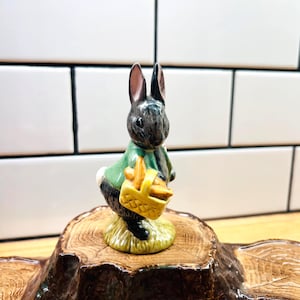 May include: A ceramic figurine of a black rabbit with long ears, wearing a green jacket and carrying a yellow woven basket filled with orange carrots. The rabbit stands on a textured, circular base painted to resemble a tree stump with grass.