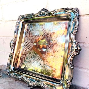 May include: A silver-colored, rectangular serving tray with ornate, scrolling edges. The tray has a reflective surface with a detailed, etched floral pattern. The tray is leaning against a white brick wall and rests on a wooden surface.