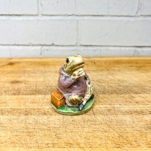 May include: A ceramic figurine of a frog wearing a mauve hooded jacket, sitting on a green base. The frog is holding a small object near its face and is positioned next to a brown rectangular basket. The figure features painted details.