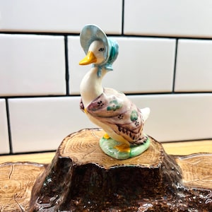 May include: A ceramic duck figurine wearing a light blue bonnet and a patterned shawl with floral accents. The duck has a yellow beak and feet, and it is standing on a small green base that is attached to a brown, textured tree stump platform.
