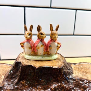 May include: A ceramic figurine featuring three brown rabbits standing side by side, each wearing a pink cape. The rabbits are positioned on a light green base, which is set atop a brown, textured, tree-stump-shaped ceramic base.