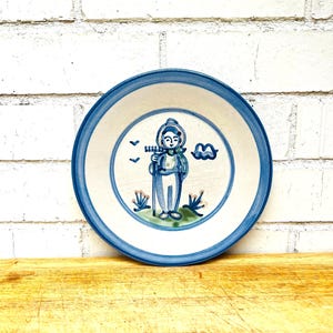 May include: A round ceramic plate with a white base and a blue rim. The center features a hand-painted blue and green illustration of a figure holding a rake, surrounded by small stylized birds, a cloud, and plants on a patch of green ground.