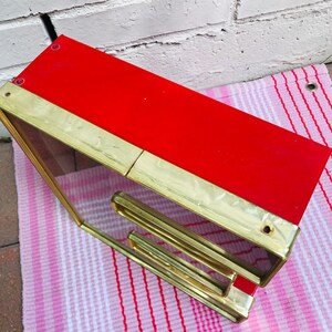 May include: A red and gold framed mirror with a decorative design. The mirror is angled and sitting on a pink and white striped surface.