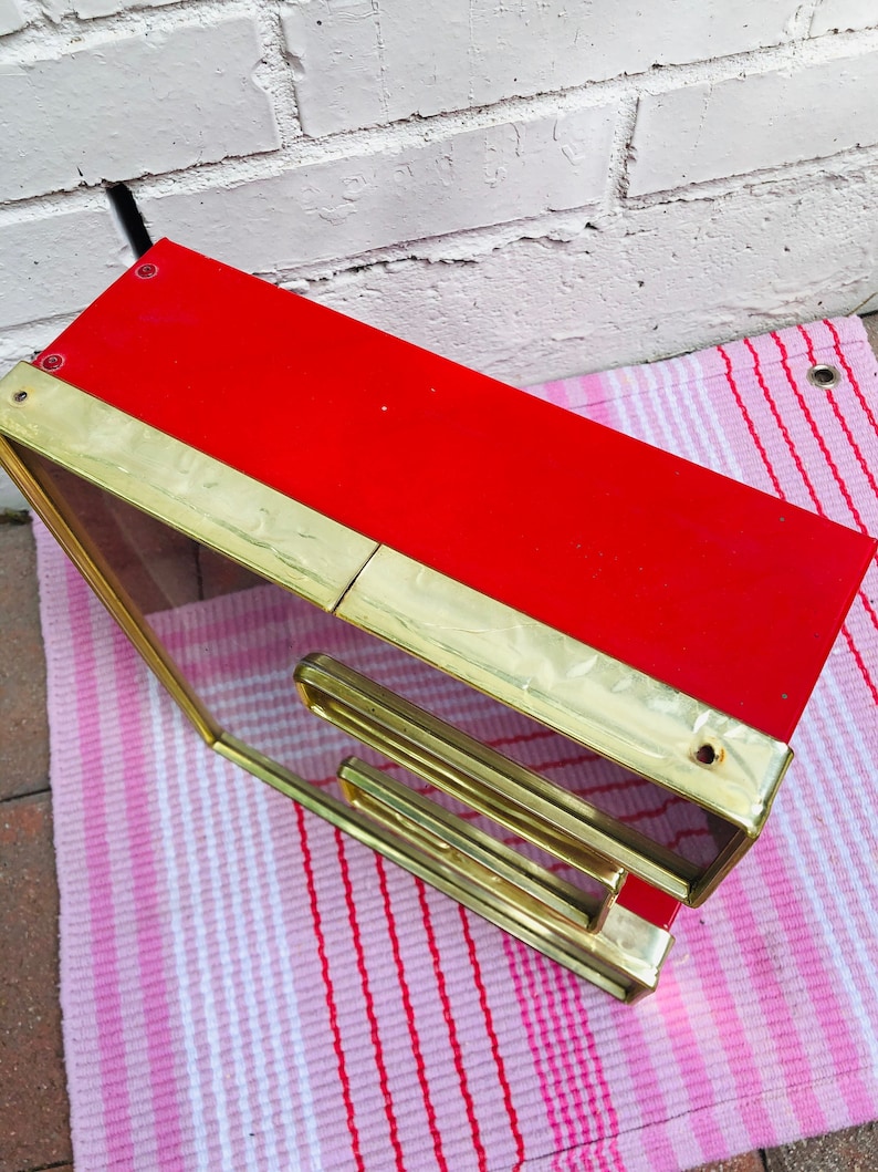 May include: A red and gold framed mirror with a decorative design. The mirror is angled and sitting on a pink and white striped surface.