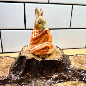 May include: A ceramic figurine of a brown rabbit wrapped in an orange blanket with white stripes, positioned on a brown ceramic base textured to resemble a tree stump with visible wood grain patterns and a glossy, dark brown finish.