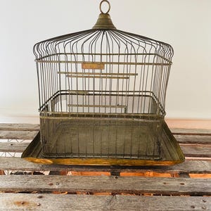 May include: An antique birdcage with a brass finish. The cage features a wire frame, a rectangular base, and a small brass tag. The cage is sitting on a wooden surface.