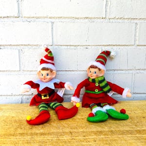 May include: Two Christmas elf dolls. Each elf is wearing a red outfit with green and white striped accents. One elf has green and white striped legs, while the other has a green and white striped scarf. Both have red hats with white trim.