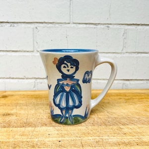 May include: A ceramic mug with a cream-colored exterior and a blue interior. The mug features a hand-painted blue illustration of a figure wearing a dress, accented with small orange and green details. It has a curved handle on the right side.