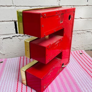 May include: A red metal letter E with gold trim. The letter is made of three sections and has a hole in the top section. The letter is sitting on a pink and white striped surface.