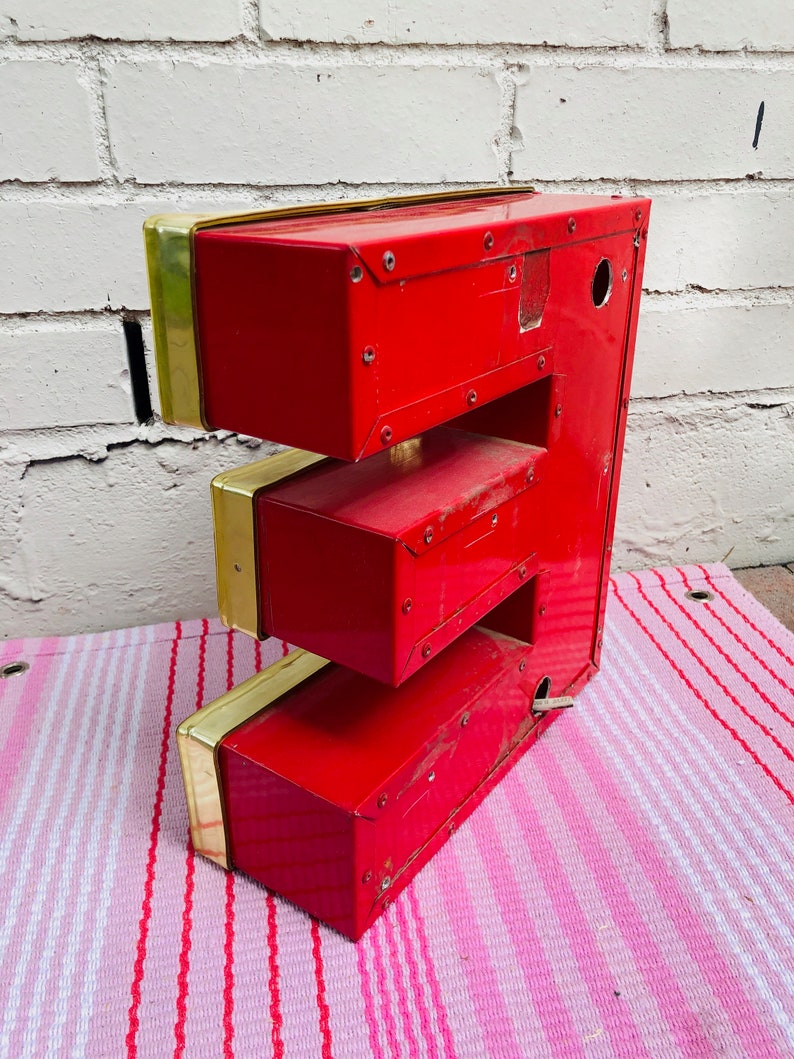 May include: A red metal letter E with gold trim. The letter is made of three sections and has a hole in the top section. The letter is sitting on a pink and white striped surface.