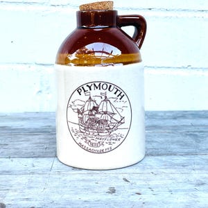 May include: A ceramic jug with a brown top and handle, a tan band, and a white base. The jug features a circular emblem with a ship illustration and the text "PLYMOUTH MASSACHUSETTS 1620 MAYFLOWER". The jug is topped with a cork.