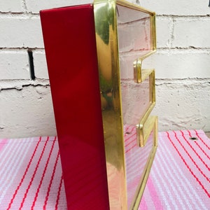 May include: A red and gold letter E sign with a clear glass front. The sign is made of metal and has a vintage look.