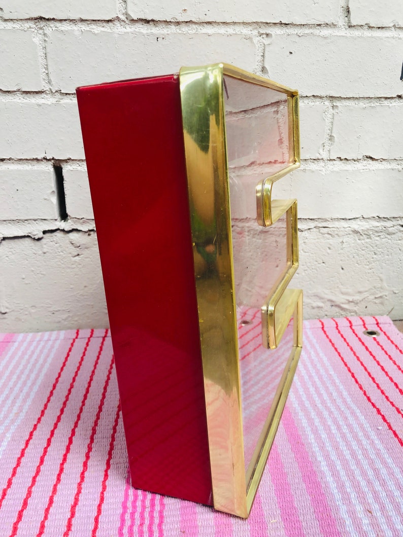 May include: A red and gold letter E sign with a clear glass front. The sign is made of metal and has a vintage look.