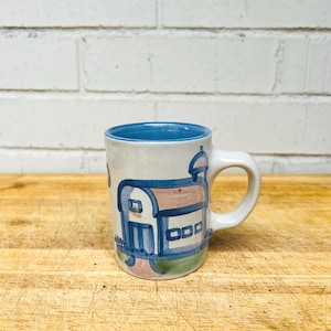 May include: A ceramic mug with a white base and a blue interior. The exterior features a hand-painted blue and brown illustration of a small house with a fence and a chimney. The mug has a curved handle on the right side and a smooth finish.