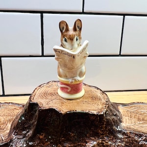 May include: A ceramic figurine of a brown mouse sitting on a yellow and pink thread spool, reading a newspaper with the text The Tailor & Cutter printed on it. The mouse has large ears, dark eyes, and a light-colored belly, perched on a base.